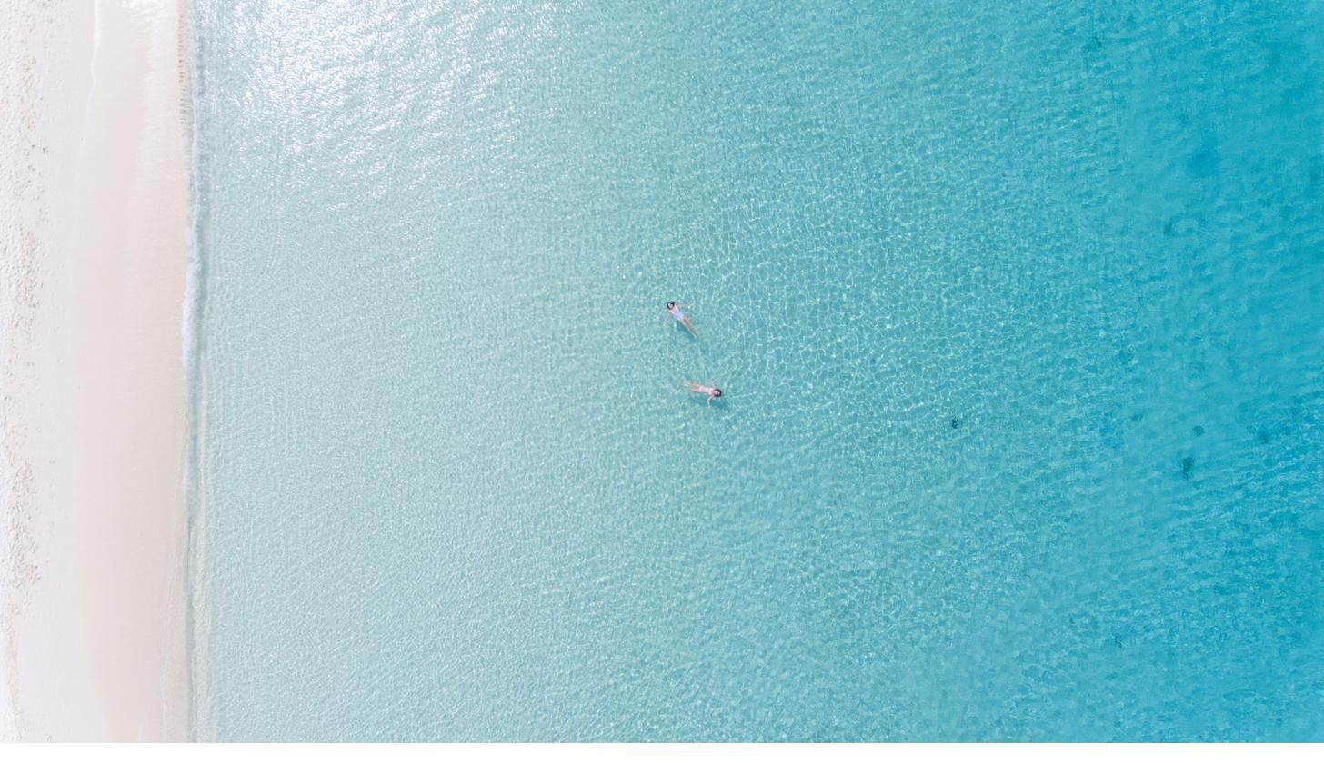 Two people floating in clear turquoise water off a Grand Cayman beach, seen from directly above